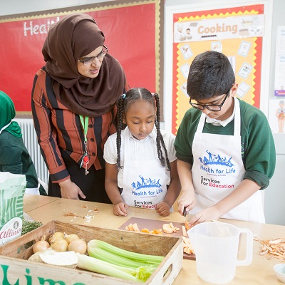 Picture of two children cooking with their teacher. Symbolises the Health For Life Programme